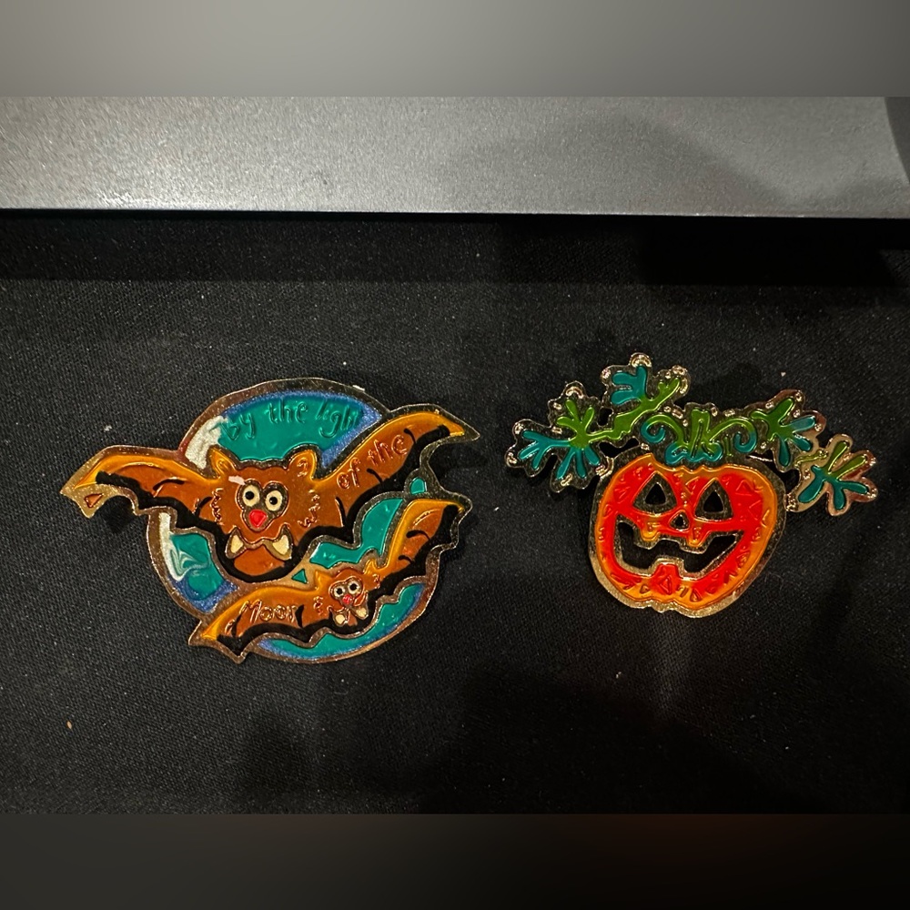 Two holiday pins by two hands, one pumpkin one that has two bats